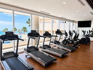 Ein Fitnessstudio mit Laufbändern und Ergometer, große Fenster mit Blick auf Palmen und das Meer.