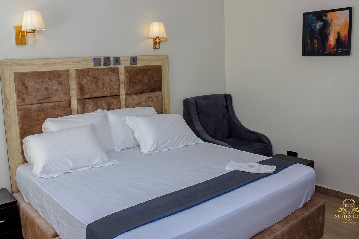 A neatly made bed with white linens, a headboard with a wooden panel design, two wall-mounted lamps, and a framed abstract painting on the wall.