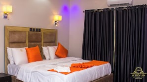 A hotel room with a bed, pillows, a headboard, wall sconces, an air conditioner, and a curtain. - P.I.O Suites (Abraka)
