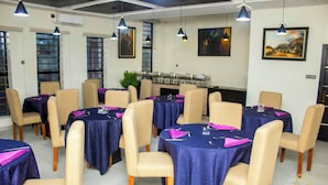 A dining area with round tables covered in blue tablecloths, beige chairs, and purple napkins. - P.I.O Suites (Abraka)