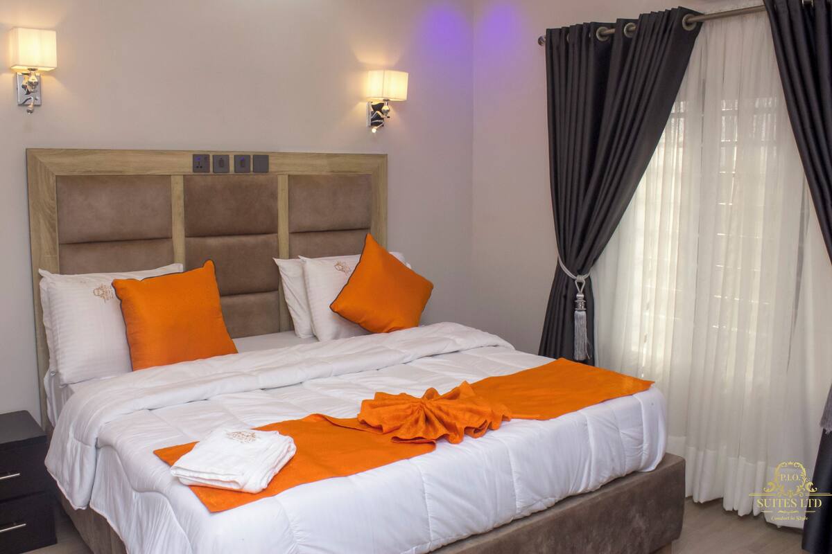 A hotel room with a large bed, two wall-mounted lamps, a wooden headboard, and a window with dark curtains.