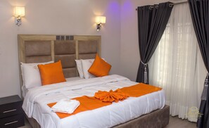 A hotel room with a large bed, two wall-mounted lamps, a wooden headboard, and a window with dark curtains.
