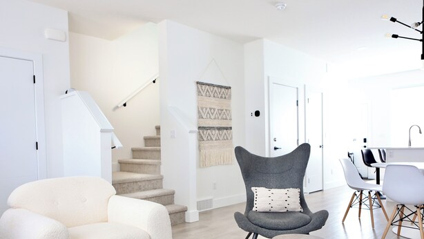 Un salon moderne avec un canapé blanc, un fauteuil gris et une petite table en bois. On aperçoit un escalier au fond, qui mène à une autre pièce.