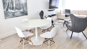 Un salon moderne avec une table de salle à manger ronde, des chaises blanches, un fauteuil gris, un téléviseur à écran plat fixé au mur et un grand tableau représentant une forêt.