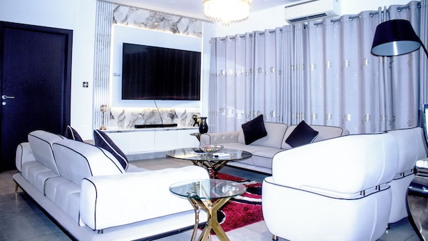 A modern living room with a sofa, armchairs, a glass coffee table, a chandelier, and a large TV. - Immaculate Beauty Gem Location Pools Gym (Accra)