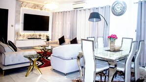 A modern living room with a sofa, a coffee table, a dining table with chairs, a TV mounted on the wall, and a clock. - Immaculate Beauty Gem Location Pools Gym (Accra)