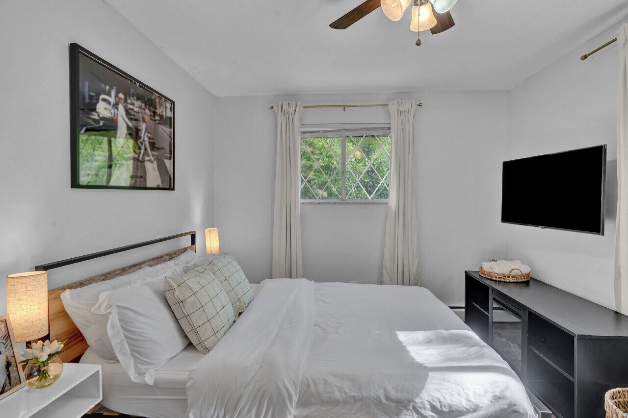 Ein Schlafzimmer mit einem Bett, einem Fernseher, einem Fenster mit Vorhängen und einem Deckenventilator.