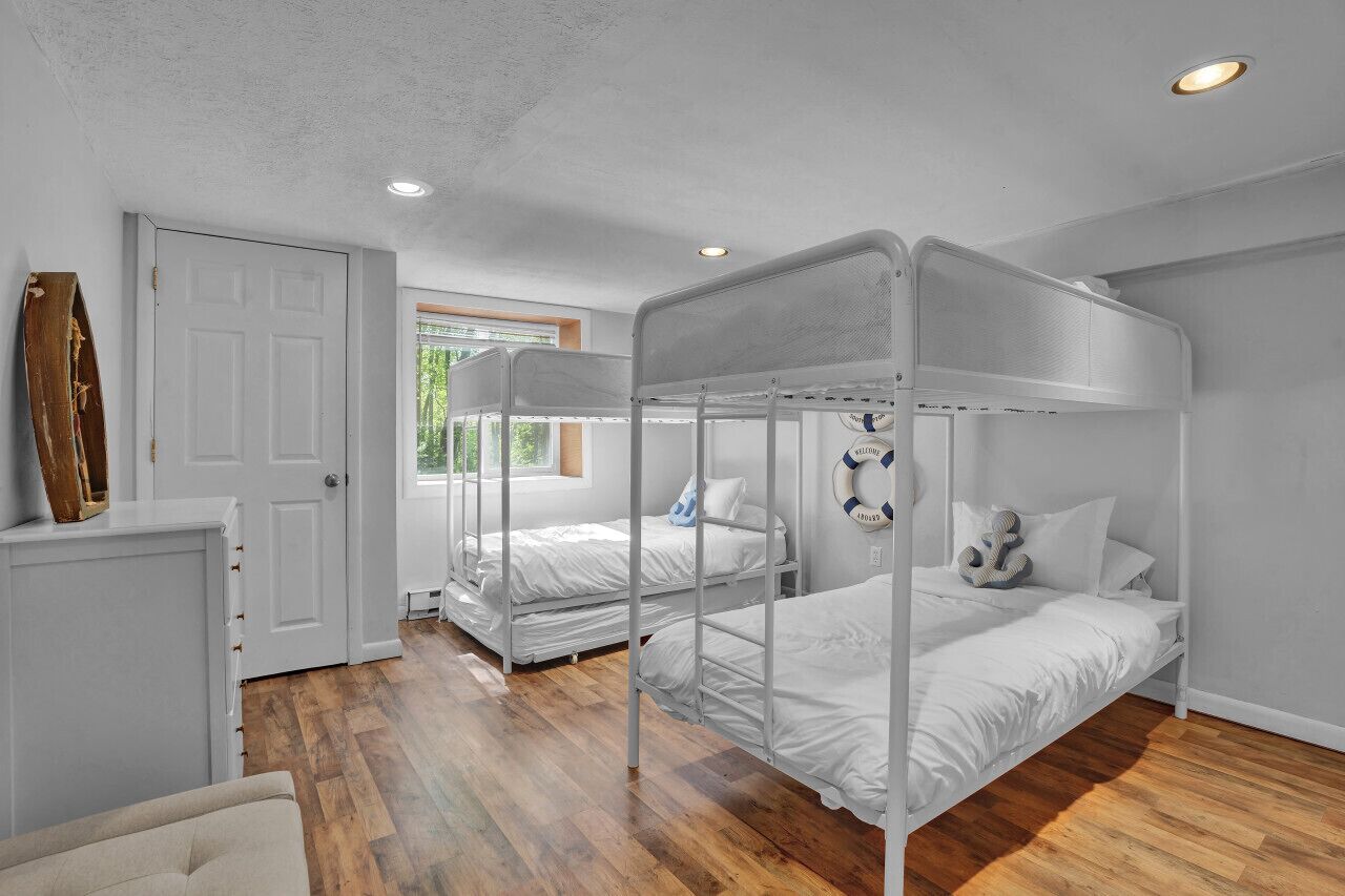 A room with bunk beds, a white door, a window with a view of greenery, and a wooden floor.
