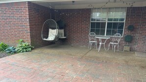 Een terras met bakstenen vloer, twee hangstoelen, een tafel met stoelen en een raam.