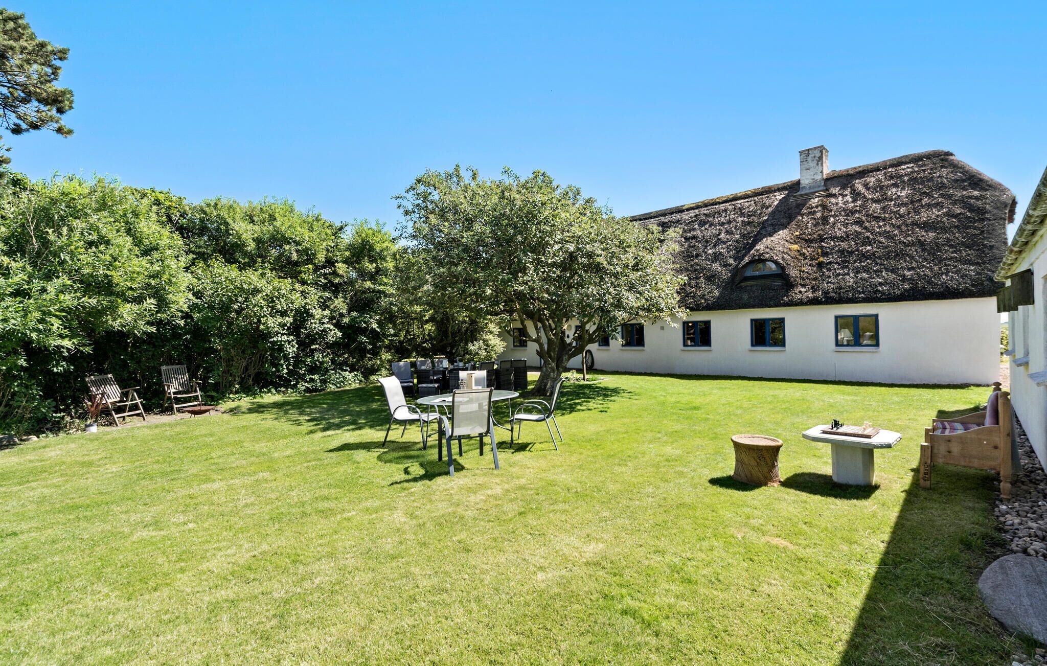A well-maintained garden with a dining area, a thatched-roof building, and a white wall with windows.