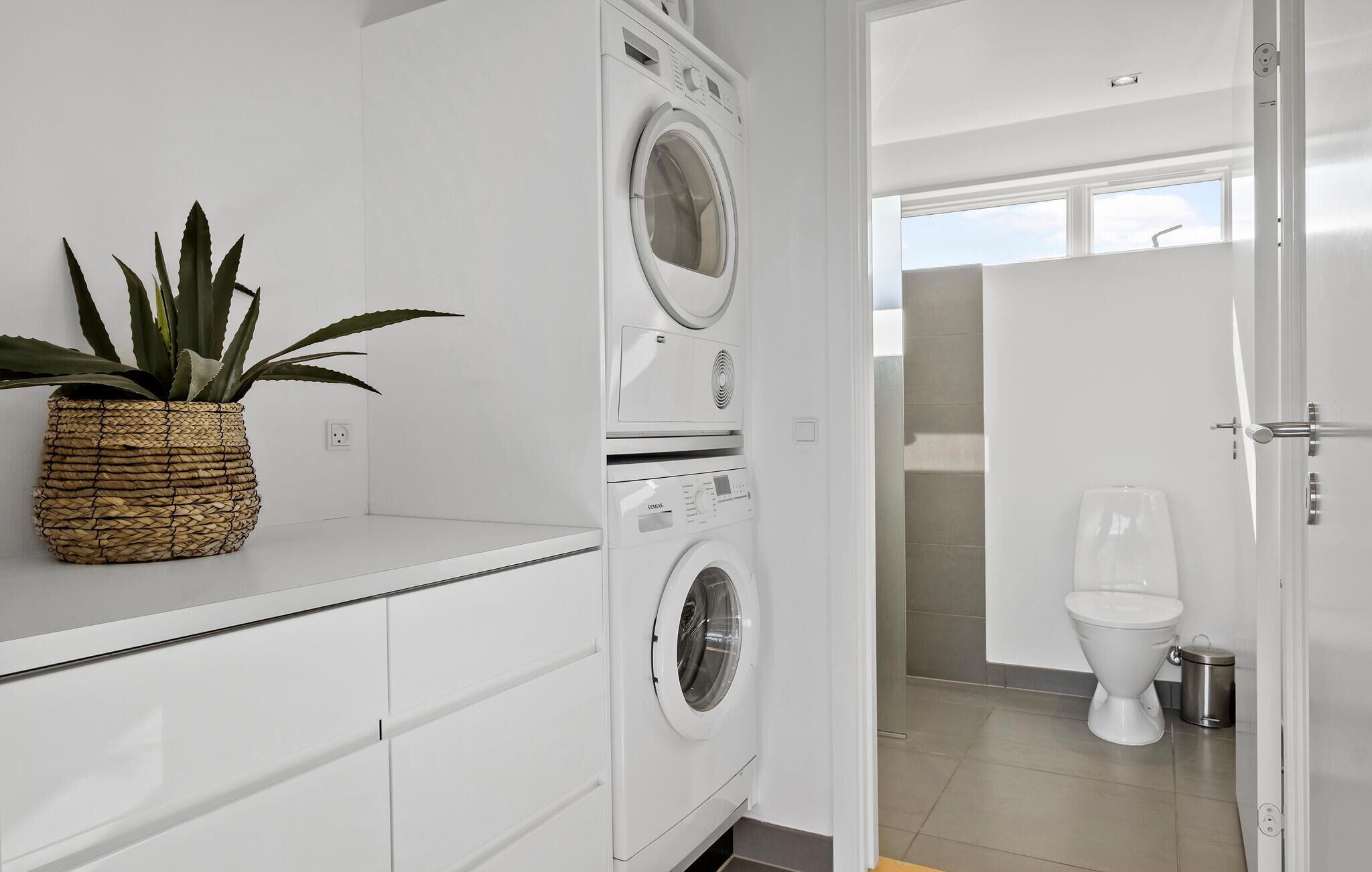 A modern laundry room with stacked washing machines, a potted plant, and a view of the bathroom.