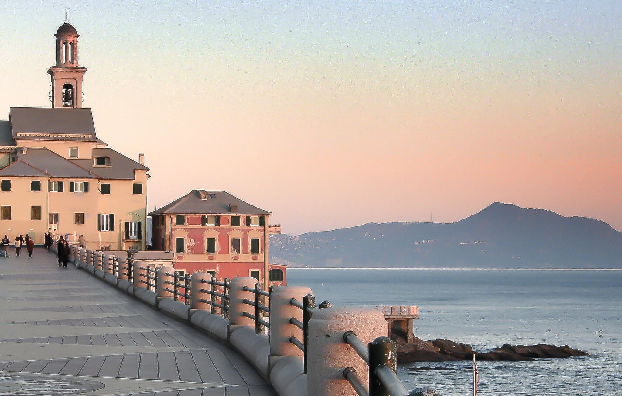 A waterfront promenade with buildings, a bell tower, and a view of the sea and mountains.