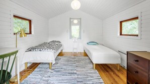 Une petite pièce aux murs recouverts de lambris blancs, comprenant deux lits, un bureau, une chaise et une commode.