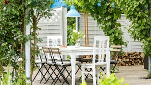 Une terrasse extérieure avec une table et des chaises blanches, une lampe suspendue bleue et une clôture blanche en arrière-plan.