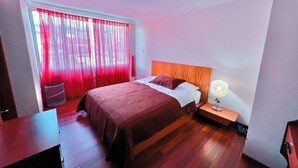 A bedroom with a bed, a nightstand, a lamp, a window with red curtains, and a trash bin. - Departamento Centrico en Loja (Loja)