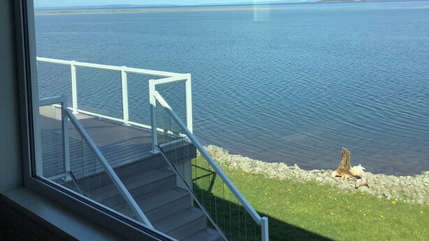 A view from a window overlooking a calm body of water, with a grassy area and a white railing leading to a deck. - Sunset Retreat Awaits (Big Pond)