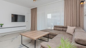 A modern living room with a sectional sofa, a wooden coffee table, a TV mounted on the wall, and a large window with sheer curtains. - Luxurious home in Vrsi (Vrsi)