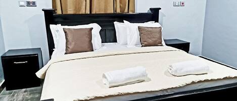 A neatly made bed with white and brown pillows, a beige bedspread, and two folded white towels.