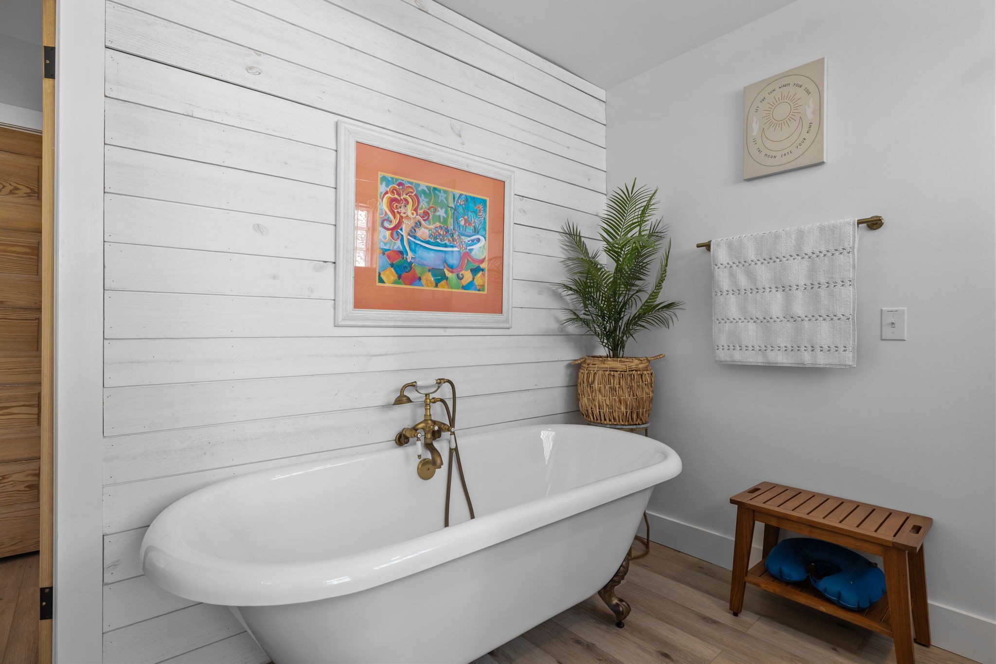 Un baño con una bañera independiente, un banco de madera, una planta en maceta y dos cuadros en la pared.