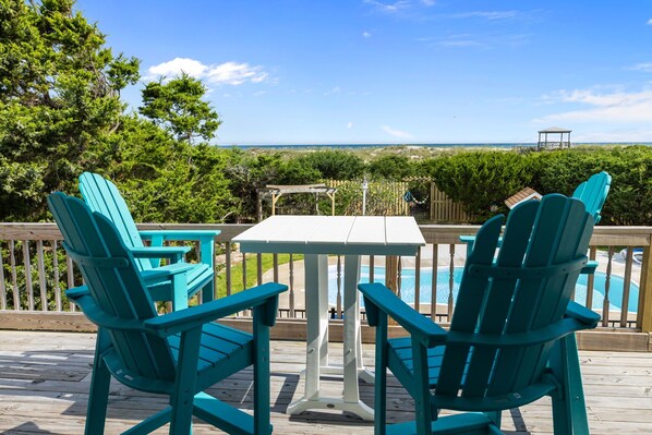A deck with turquoise Adirondack chairs and a white table, overlooking a pool and a beach.