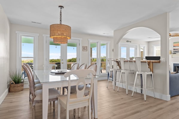 A spacious dining area with a large table and chairs, a hanging light fixture, and a view of the outdoors through large windows.