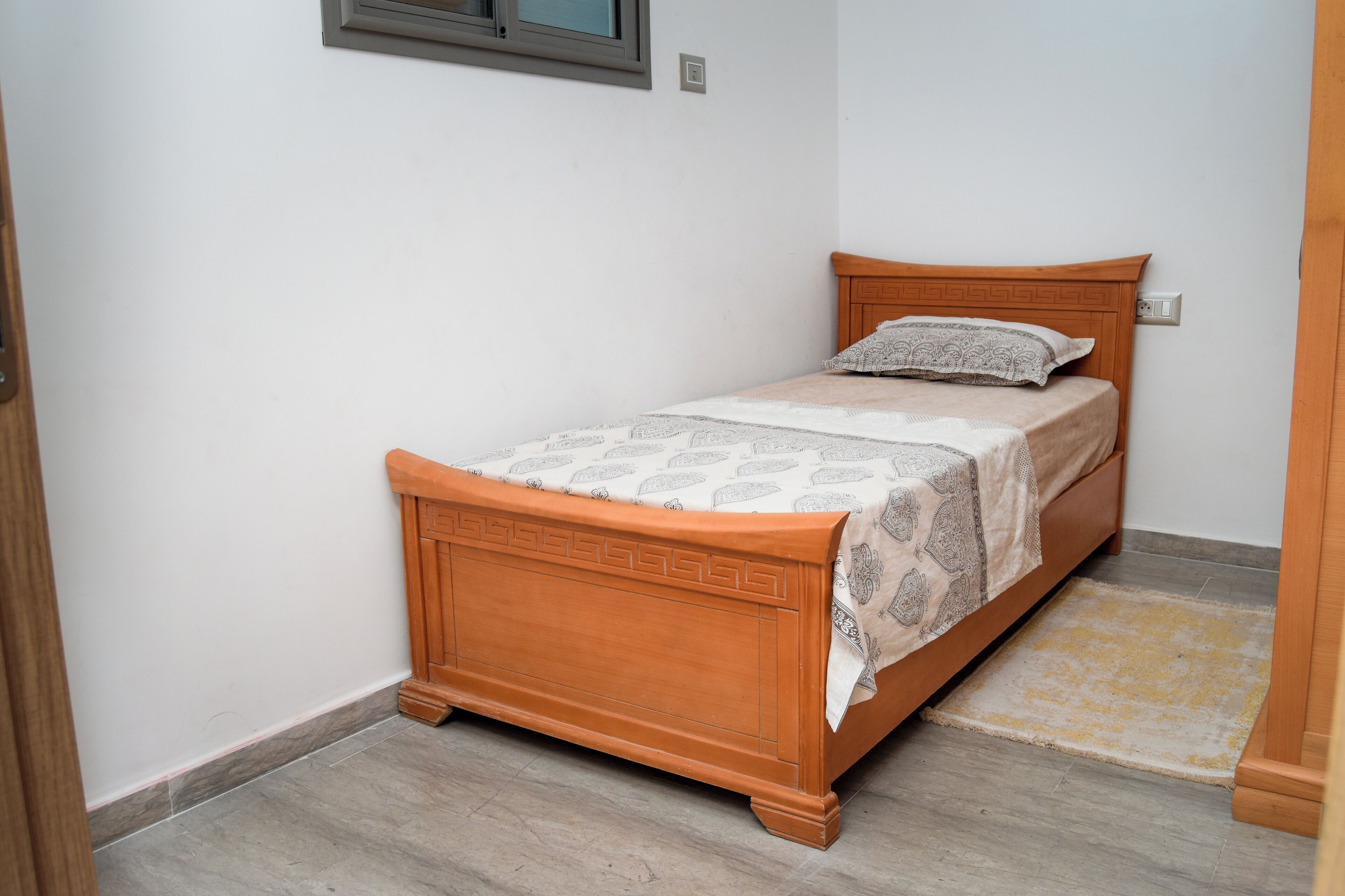 Ein Einzelbett mit Holz Kopfteil und einem gemusterten Bettbezug, an einer weißen Wand mit einem kleinen Fenster darüber platziert.