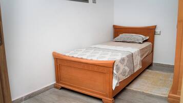 A single bed with a wooden headboard and a patterned bedspread, positioned against a white wall with a small window above it.