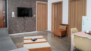 A modern living room with a flat-screen TV mounted on the wall, a sofa, a coffee table, and a dining area with a table and chairs.