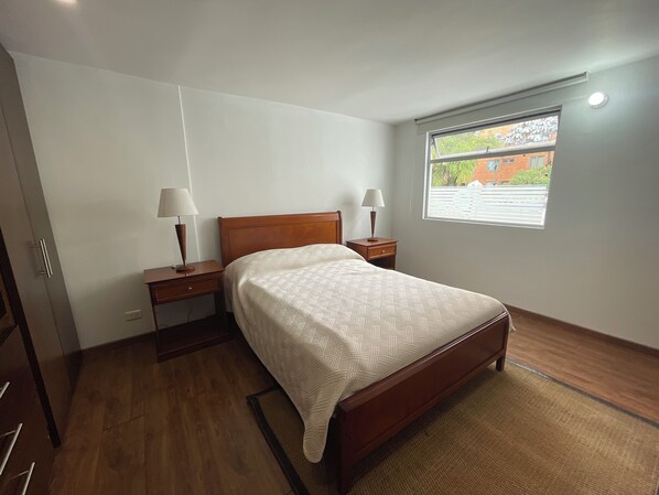A bedroom with a wooden bed, two bedside tables with lamps, a window with blinds, and a view of the outside. - Súper Apartamento TV Wi Fi y parqueadero (Bogotá)