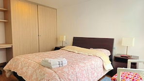 Uma cama bem arrumada com edredom floral rosa e uma toalha branca dobrada ao lado.