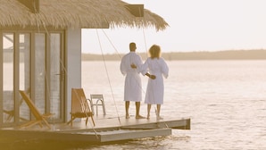 A couple standing on a wooden deck with a thatched roof, overlooking a calm body of water. - Premium Floating Bugalow by Stay Ombi in the Keys with Stunning Ocean Views. (Key Largo)