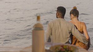 A couple sitting at a wooden table by the sea, with a bottle and a plate of food in front of them. - Premium Floating Bugalow by Stay Ombi in the Keys with Stunning Ocean Views. (Key Largo)