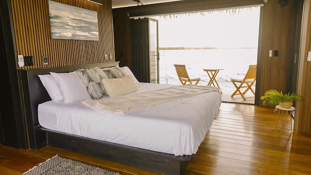 A bedroom with a large bed, wooden flooring, a view of the water, and a balcony with chairs. - Premium Floating Bugalow by Stay Ombi in the Keys with Stunning Ocean Views. (Key Largo)