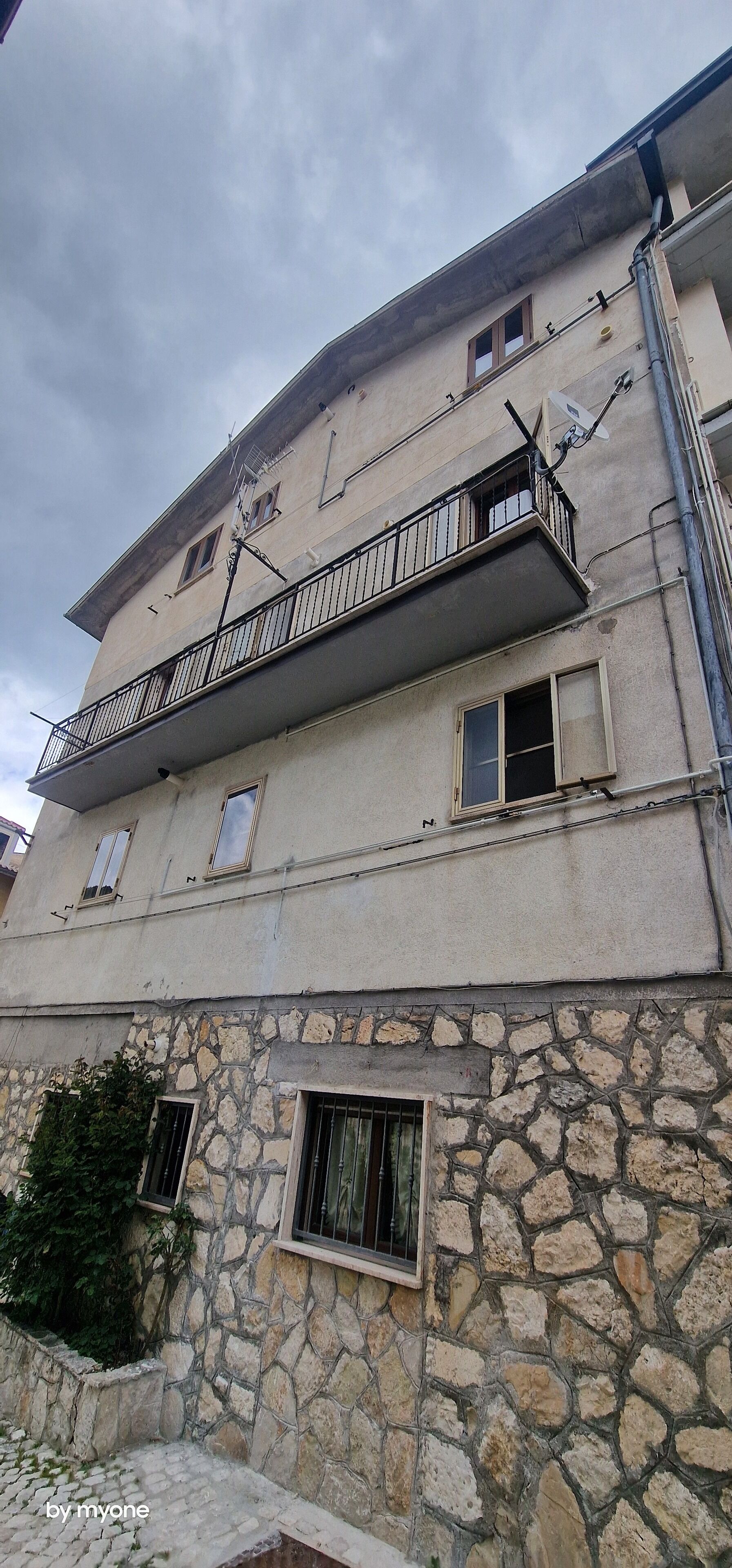 A multi-story building with balconies, stone walls, and a flat roof.