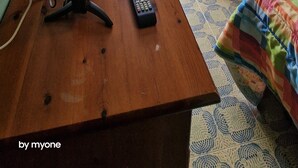 Een kamer met een houten bureau, een computer beeldscherm en een stoel. Er is een raam met gordijnen, een houten kledingkast en een bed met een kleurrijke deken.