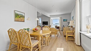 Un coin repas avec des chaises en osier et une table ronde, un téléviseur et une vue sur la mer grâce à de grandes fenêtres.