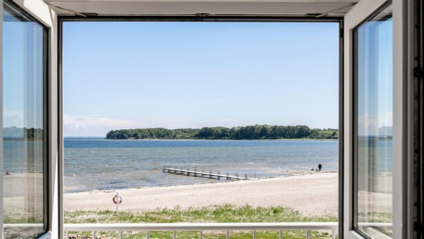 Une vue depuis une fenêtre donnant sur une plage, avec une jetée s’avançant dans l’eau, une bouée de sauvetage sur le sable et une île boisée à l’horizon.