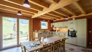 A spacious kitchen with wooden beams, a dining area with a table and chairs, and a view of a grassy outdoor area. - Apartment 'Le Chalet De La Source' with Mountain View, Private Terrace and Wi-Fi (Montagnole)