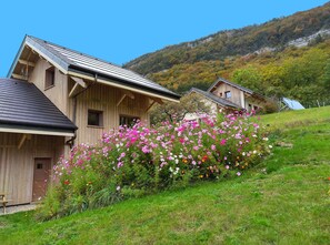 A wooden cabin with a sloped roof, surrounded by a garden of colorful flowers, set against a hillside. - Apartment 'Le Chalet De La Source' with Mountain View, Private Terrace and Wi-Fi (Montagnole)