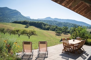 Une terrasse extérieure avec une table et des chaises, offrant une vue sur un paysage vallonné, parsemé d’arbres, sous un ciel dégagé.