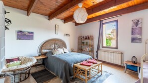 Une chambre confortable avec un plafond en bois, un lit recouvert d’une couverture bleue, une chaise en osier, une table de chevet, un radiateur et une fenêtre donnant sur un beau paysage.