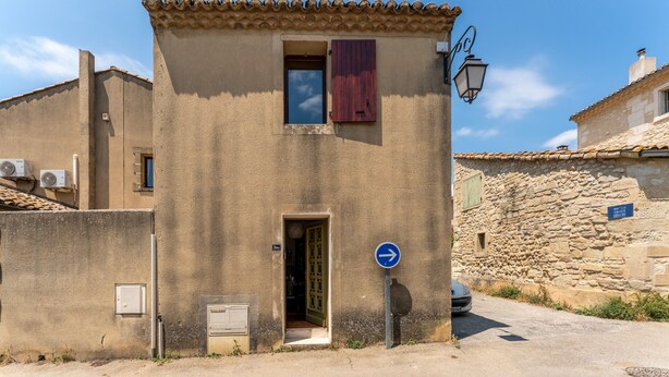 Un bâtiment avec une porte, une fenêtre avec des volets, un panneau de signalisation bleu indiquant la direction, et une voiture garée à proximité.