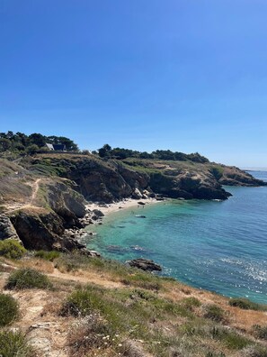 Paesaggio costiero con un sentiero che conduce a una spiaggia, terreno roccioso e vista sul mare.