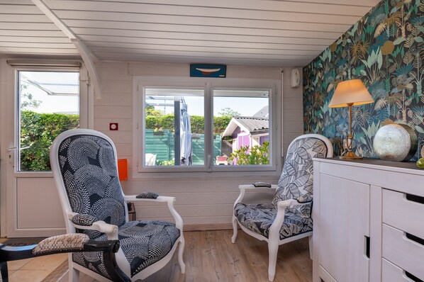 A room with two patterned chairs, a white cabinet, a table, and a window with a view of greenery and a building.