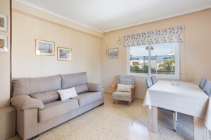 A living room with a sofa, armchair, and dining table, a window with a view, and wall-mounted pictures.