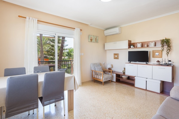 A living room with a dining table, chairs, a TV, a sofa, and a balcony with a view of palm trees.