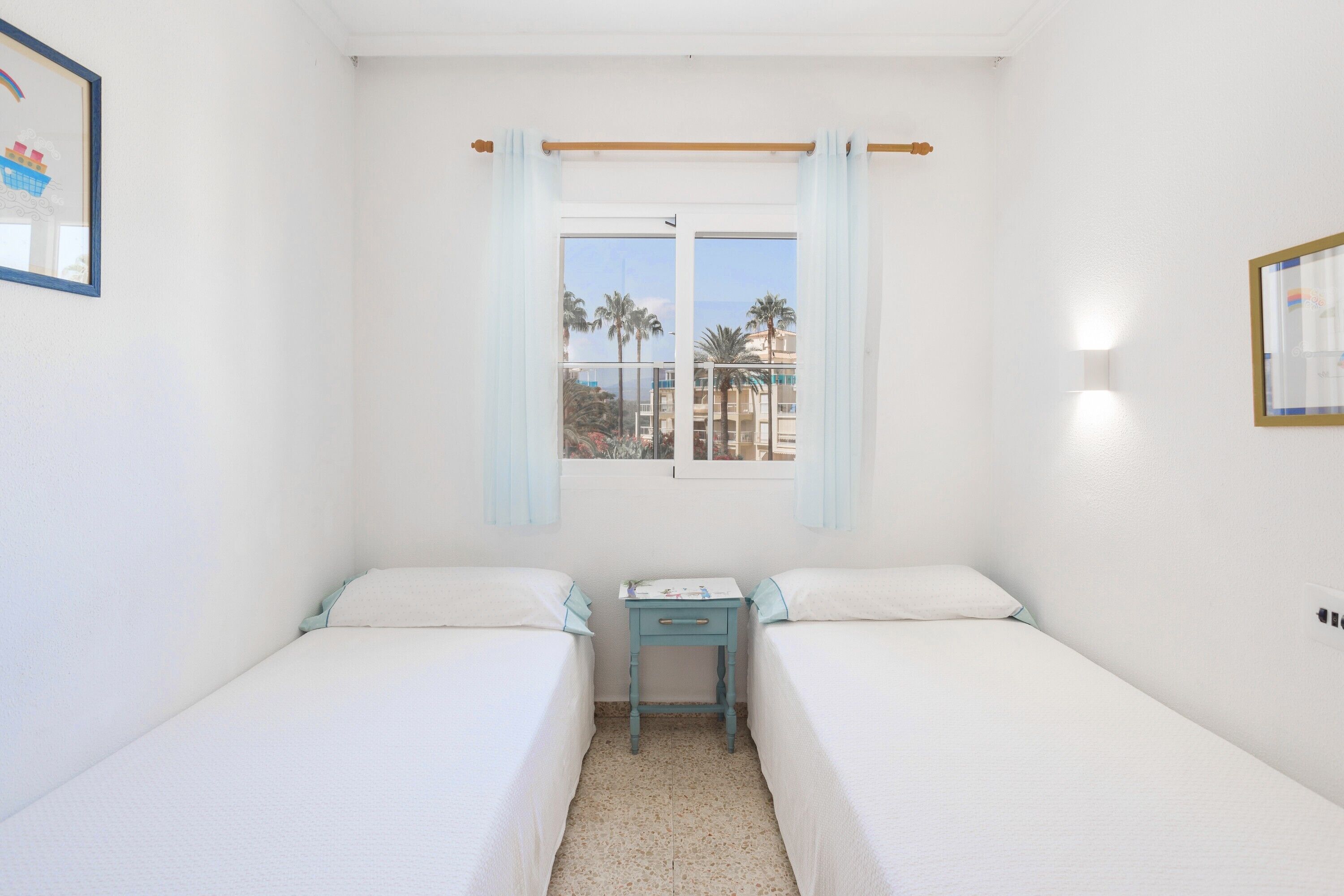 A room with two beds, a small table, a window with curtains, and a view of palm trees and buildings.