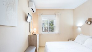 A bedroom with a bed, a bedside table with a lamp, a bench, a window with curtains, and a wall-mounted air conditioner.