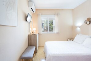 Un dormitorio con cama, una mesita de noche con lámpara, un banco, una ventana con cortinas y un aire acondicionado de pared.