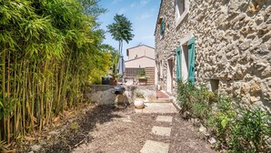Un étroit sentier de pierres mène à un bâtiment rustique, avec un jardin d’un côté et une bambouseraie de l’autre.